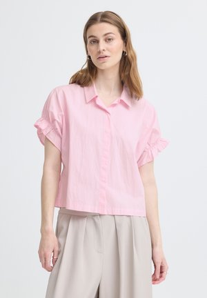 Mladá žena svetlohnedými vlasmi, ktorá má na sebe voľnú ružovú blúzku s krátkym rukávom a béžové plisované nohavice s vysokým pásom na bielom pozadí.