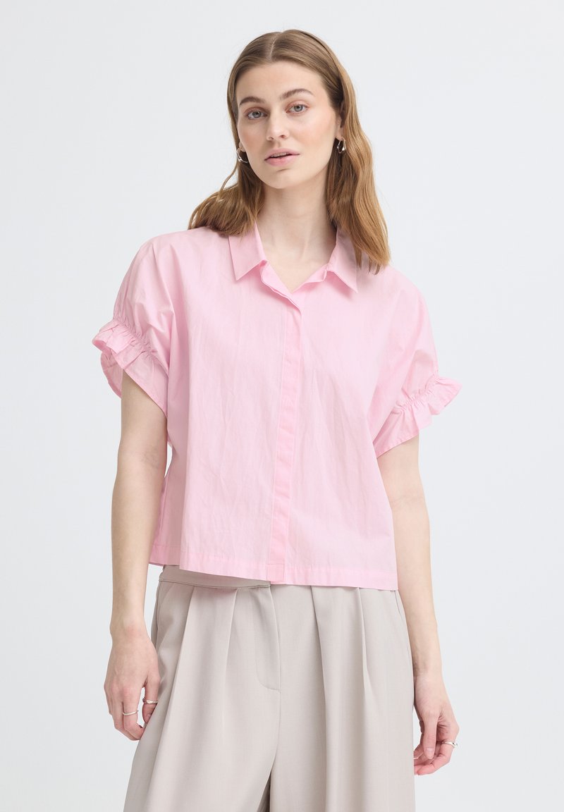 Jeune femme aux cheveux châtain clair portant un chemisier rose ample à manches courtes et un pantalon plissé beige taille haute sur fond blanc.