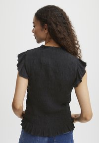 Haut noir sans manches en tissu texturé, avec des accents de manches volantés et un ourlet évasé. La vue arrière met en avant le design smocké.
