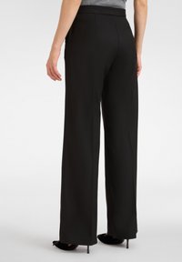 Pantaloni neri a gamba larga con una texture liscia, dotati di vita alta e tasche laterali, abbinati a tacchi neri.