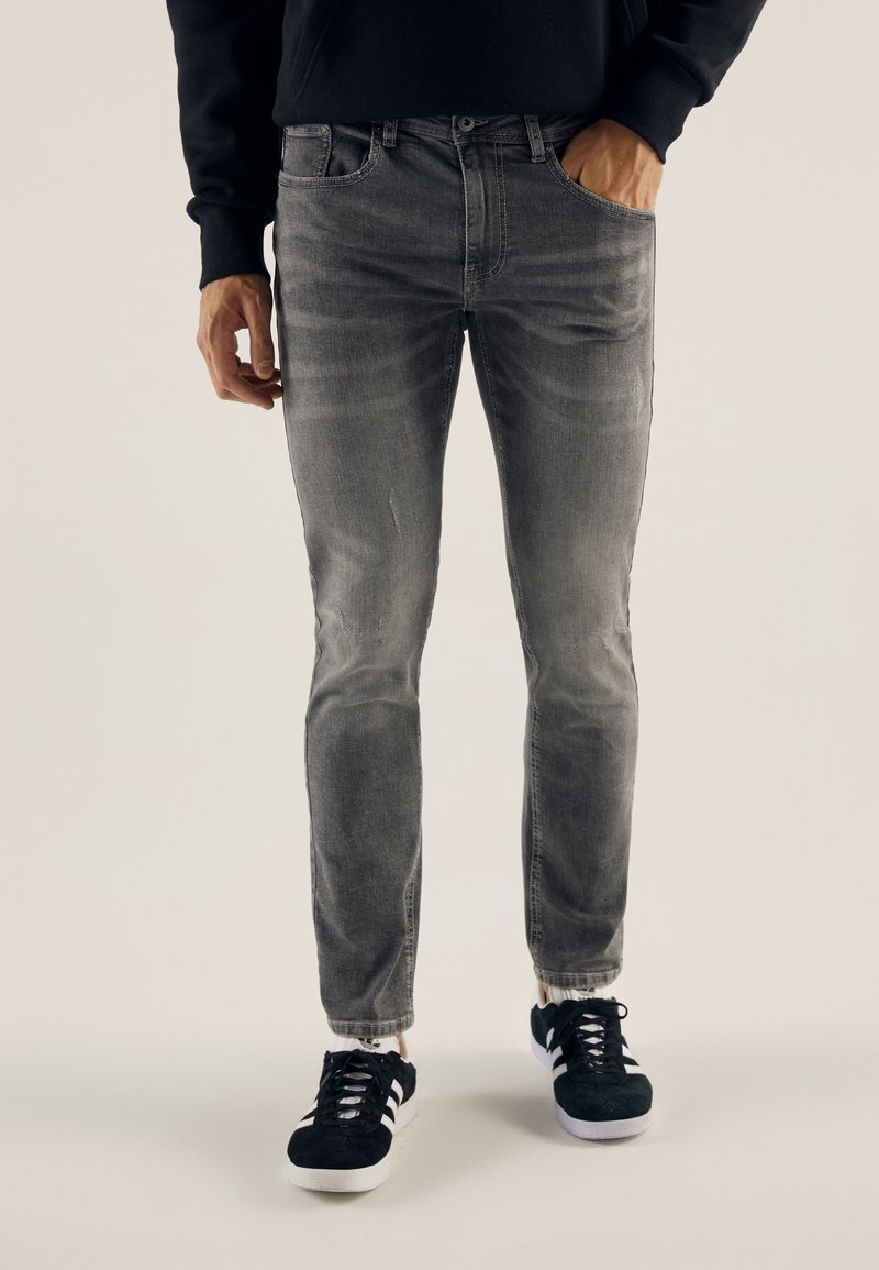 Homme portant un jean slim gris délavé et des baskets noires à rayures blanches, debout devant un fond uni.