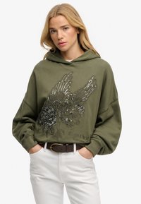Sudadera con capucha verde oliva con un diseño de pájaro con lentejuelas en el frente, con un ajuste relajado y puños acanalados, combinada con pantalones blancos y un cinturón marrón.