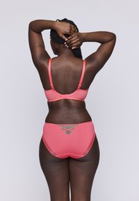 Korallivärinen bikinitsetti, jossa on texturoitu hihndesign, säädettävät hihnat ja leikkauskohtaa alaosien takana. Sujuva kangas.