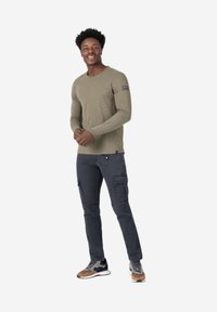 Olijfgroen shirt met lange mouwen en een ronde halslijn, met een patch op de linker mouw, gecombineerd met donkere cargobroeken en meerkleurige sneakers.