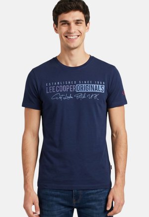 Homme souriant portant un t-shirt bleu marine avec le texte « Lee Cooper Originals » et un jean bleu foncé, debout devant un fond blanc uni.