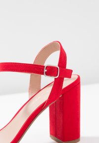 Red suede high-heeled sandal with a square block heel, adjustable ankle strap, and smooth beige insole. Features a silver buckle for closure.