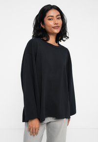 Μαύρο κοντομάνικο πουκάμισο από απαλό ύφασμα, με χαλαρή εφαρμογή και στρογγυλή λαιμόκοψη, συνδυασμένο με ανοιχτό γκρί τζιν παντελόνι.