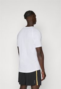 Camiseta deportiva blanca de manga corta con cuello redondo, detalles de costura en contraste y aberturas laterales, combinada con pantalones cortos negros con un acento amarillo.