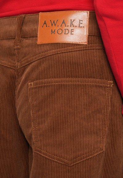 Pantalones de pana marrón con una etiqueta de cuero que dice "A.W.A.K.E. MODE." Presentan un rigido vertical y un bolsillo trasero con costura curva.
