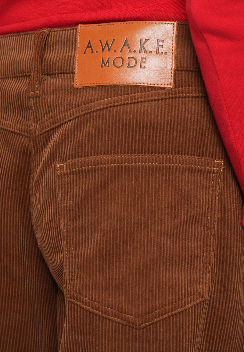 Brown corduroy pants with a leather label reading "A.W.A.K.E. MODE." Features vertical ribbing and a back pocket with curved stitching.