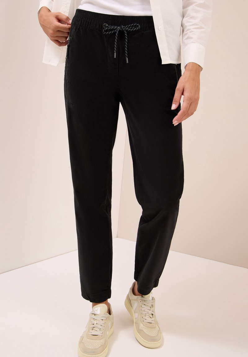 Personne portant un pantalon noir à cordon et des baskets beige, debout contre un mur et un sol de couleur claire.
