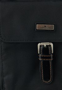 Zwarte stoffen schoudertas met een gestructureerd oppervlak, voorzien van een metalen logo plaat en een leren schouderband met contrasterende stiksels.