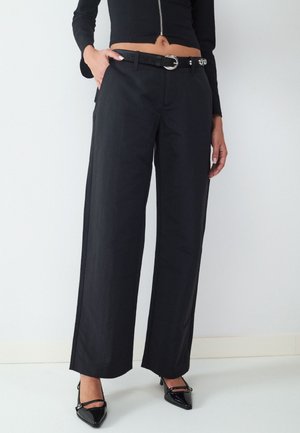 Frau trägt schwarze Hose mit weitem Bein und schwarzem Gürtel, schwarzes Zip-up cropped Top und schwarze spitze High Heels, steht vor weißem Hintergrund.