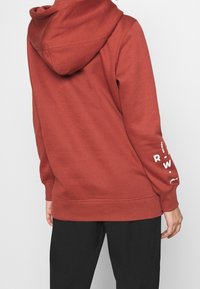 Bordeauxfärgad huvtröja i mjukt tyg, med en enkel rygg, ribbade ärmslut och vit tryckt text på högra ärmen.