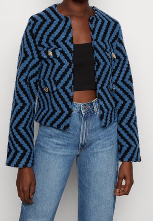 Person trägt eine blau-schwarze Jacke mit Zickzack-Struktur und goldenen Knöpfen, ein schwarzes Croptop und hellblaue Jeans mit hoher Taille.