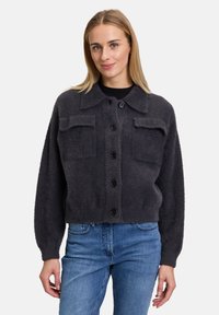 Cardigan grigio e soffice con colletto, bottoni frontali e due tasche sul petto. Taglio largo con polsini a coste. Indossato sopra una maglietta nera e jeans blu.