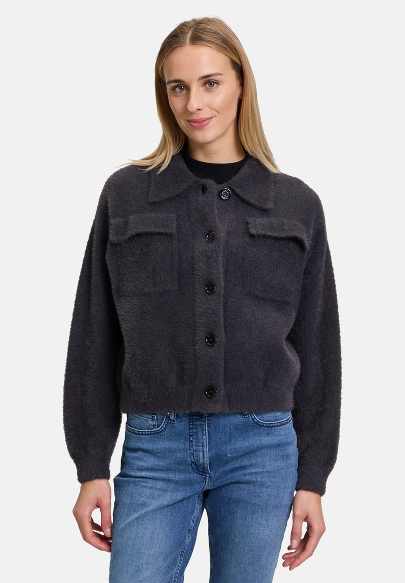 Cardigan grigio e soffice con colletto, bottoni frontali e due tasche sul petto. Taglio largo con polsini a coste. Indossato sopra una maglietta nera e jeans blu.