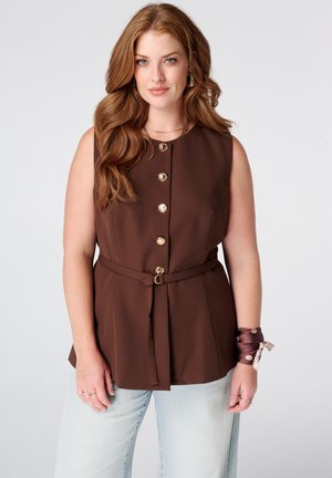 Femme aux longs cheveux roux portant un haut marron sans manches boutonné avec une ceinture à la taille et un jean bleu clair sur un fond blanc uni.