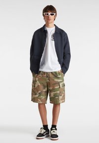 Marineblauwe button-up jas, wit graphic t-shirt, camo cargo shorts en zwarte Vans schoenen met witte accenten, met witte zonnebril.
