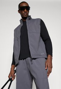 Gray vest with a high collar and zippered pockets, layered over a black long-sleeve top. Model holds a black walking stick.
