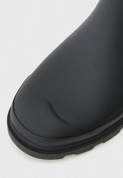 Botte en caoutchouc noire avec une texture lisse, un bout arrondi et un bord légèrement surélevé autour de la semelle. Design durable adapté aux conditions humides.