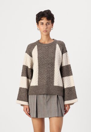 Braun- und cremefarbener Kabelstrickpullover mit weiten Ärmeln, der abwechselnde Streifen und einen runden Ausschnitt aufweist. Kombiniert mit einem plissierten Karorock.