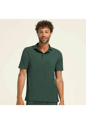 Uomo con capelli ricci che indossa una polo a maniche corte verde scuro, in piedi davanti a uno sfondo chiaro semplice, con tatuaggi visibili sugli avambracci.