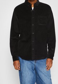 Camicia di velluto a coste nera con colletto a bottoni, tasca frontale e maniche lunghe, abbinata a pantaloni in denim blu. Tessuto texture visibile.