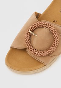Beige Slidesandale aus weichem Wildleder mit großem, rundem Akzent aus kleinen beigen Perlen und einem goldenen Stift. Strukturiertes Fußbett für Komfort.