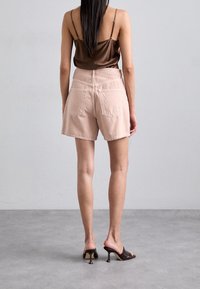 Shorts de mezclilla rosa claro con un corte relajado, que cuentan con dos bolsillos traseros y un dobladillo deshilachado, combinados con una camisola de satén marrón y mule de tacón alto.