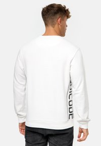 Sudadera blanca con cuello redondo y manga larga, con un logo impreso en negro en el costado, fabricada en suave algodón.
