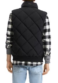 Schwarze, gesteppte Puffer-Weste mit hohem Kragen, mit einem Logo am Nacken, getragen über einem schwarz-weißen karierten Hemd. Hellblaue Jeans.