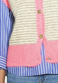 Pruhovaný béžový a bílý pletený cardigan s jasně růžovou spodní částí a knoflíkovým zapínáním, vrstvený přes modro-bílou pruhovanou košili.