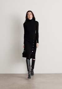 Čierne pletené šaty s vysokým golierom, bočným rozparkom a detailmi na gombíky. Modelka má na sebe čižmy po kolená a drží malú tašku.