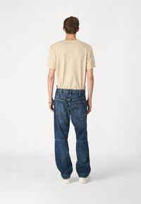 Samanis medvilninis T-shirt ir laisvi mėlyni džinsai. Džinsai turi du užpakalinius kišenių su išlenktomis siūlėmis. Modelis dėvi baltus sportbačius.