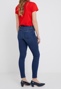 Jeans ajustados de mezclilla azul oscuro, con una cintura de media altura, bolsillos traseros y una textura suave. Combinados con una blusa corta de manga corta roja y zapatos oscuros.