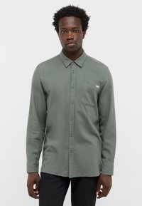 Zielona koszula zapinana na guziki z kołnierzykiem, kieszenią na piersi i długimi rękawami. Materiał ma teksturowane wykończenie i lekko luźny krój.