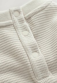 Tissu texturé blanc cassé avec un motif en nid d'abeille, présentant une rangée de boutons circulaires de teinte claire au centre.