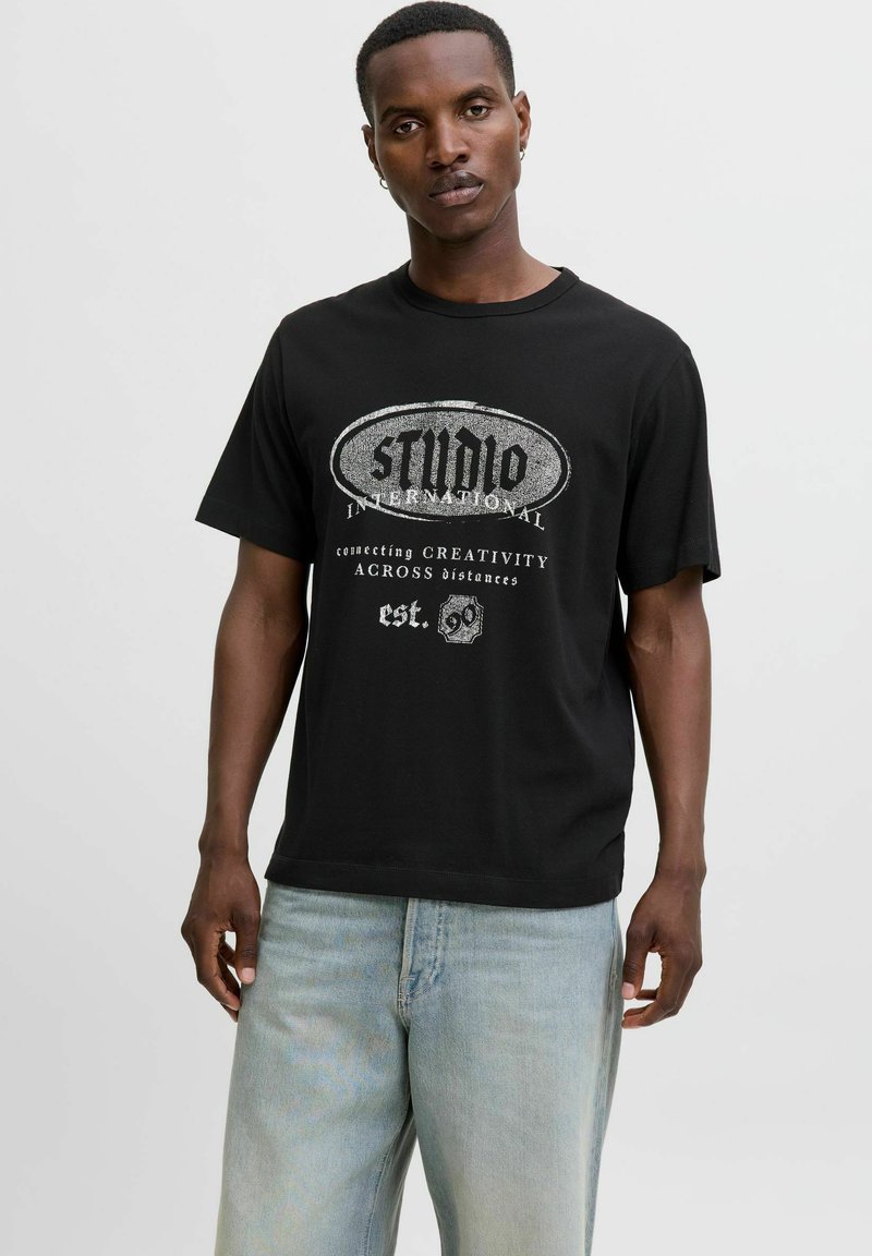 Camiseta de algodón negra con un diseño gráfico blanco que incluye texto, cuello redondo, mangas cortas y un corte relajado, combinada con jeans de color azul claro.