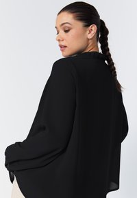 Black blouse with long sleeves, lightweight fabric, loose fit, pleated detailing at the back, and buttoned cuffs.