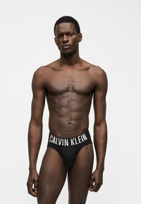 Μυώδης άνδρας που φοράει μαύρα εσώρουχα Calvin Klein, στέκεται μπροστά σε απλό ανοιχτό γκρι φόντο, κοιτάζοντας μπροστά με ουδέτερη έκφραση.