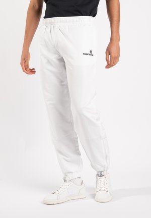 Person iført hvide joggers med elastisk talje og ankler med Sergio Tacchini-logo, kombineret med hvide sneakers og en sort top.