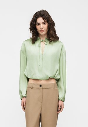 Femme aux cheveux bouclés portant un chemisier vert clair à manches longues avec un col noué et un pantalon taille haute beige sur un fond uni.