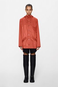 Αδιάβροχο κόκκινο μπουφάν με κουκούλα, κουμπιά με τσαμπί και δύο μπροστινές τσέπες, φορεμένο πάνω από μαύρες σορτς και ψηλές μαύρες λαστιχένιες μπότες. Λείος υφής.