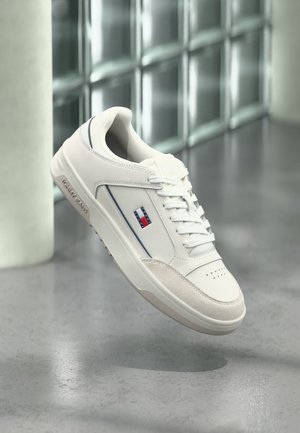 Baskets blanches Tommy Jeans avec logo sur le côté, lacets blancs et semelle en caoutchouc, présentées sur un fond intérieur moderne flou.