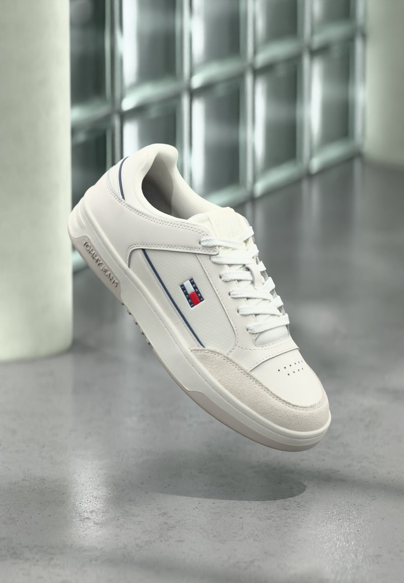 Baskets blanches Tommy Jeans avec logo sur le côté, lacets blancs et semelle en caoutchouc, présentées sur un fond intérieur moderne flou.