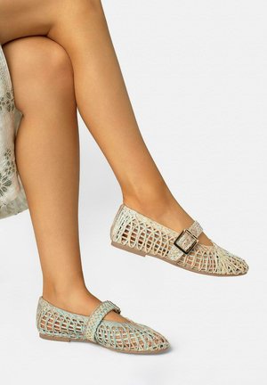 Gekreuzte Beine, die beige flache Schuhe mit offenem Gewebe und Schnallriemen tragen, kombiniert mit einem Rock mit Blumenmuster vor einem einfarbigen Hintergrund.
