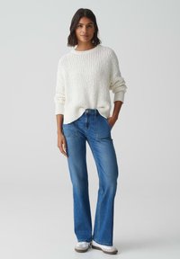 Witte, gestructureerde gebreide trui met een losse pasvorm; hoge taille blauwe jeans met een flared pijp; witte schoenen met grijze accenten.