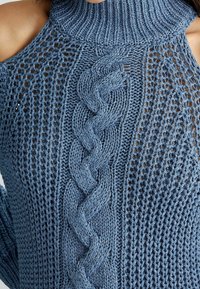 Pull en maille bleu avec col montant côtelé, épaules ouvertes, détails tressés et motif texturé à tissage ouvert.