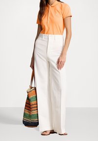 Orange polo tröja med knappar, tillsammans med vita vida byxor. Bär en vävd, flerfärgad randig tote-väska. Bruna sandaler.